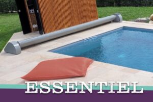 Piscine avec volet roulant automatique gris enroulé dans un abri en bois moderne. Coussin terracotta sur la plage. Texte ESSENTIEL.