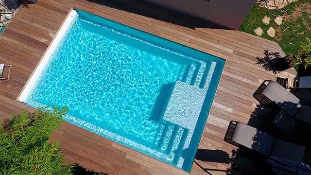 Piscine rectangulaire à l'eau bleue scintillante, intégrée dans une grande terrasse en bois. L'escalier et le banc immergés sont visibles.